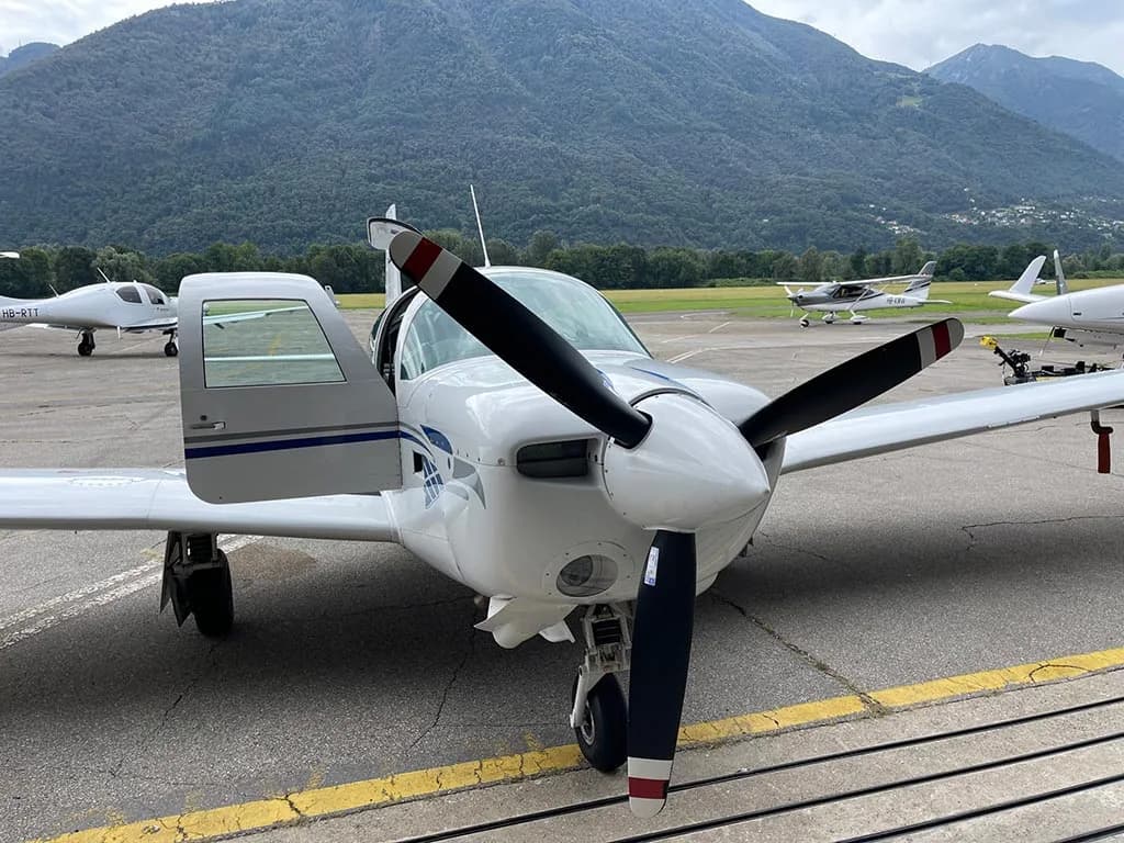 Front view of a white single-engine propeller aircraft with blue stripes, parked on an airfield with its door open and mountains in the background.