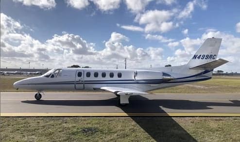 A white Cessna Citation business jet with a blue stripe is parked on an airport tarmac under a partly cloudy sky.