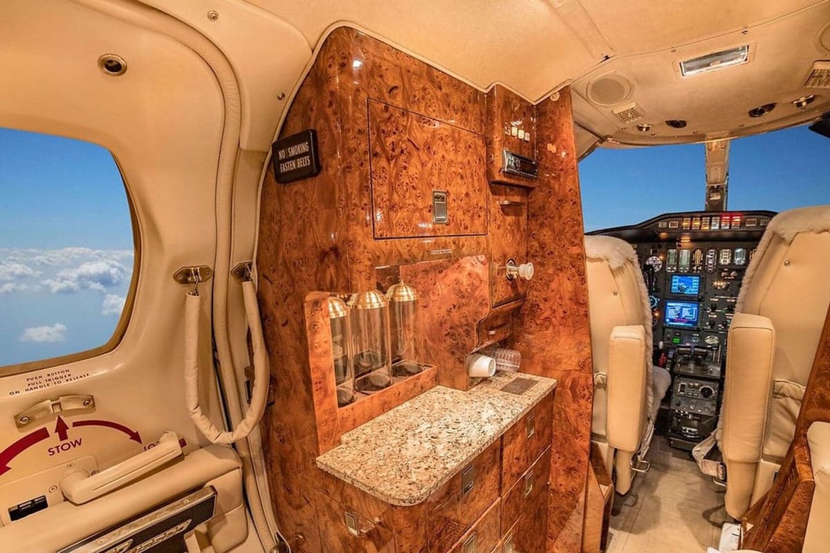 A view inside a private jet's cabin showing a galley area with polished wood paneling and a glimpse of the cockpit.