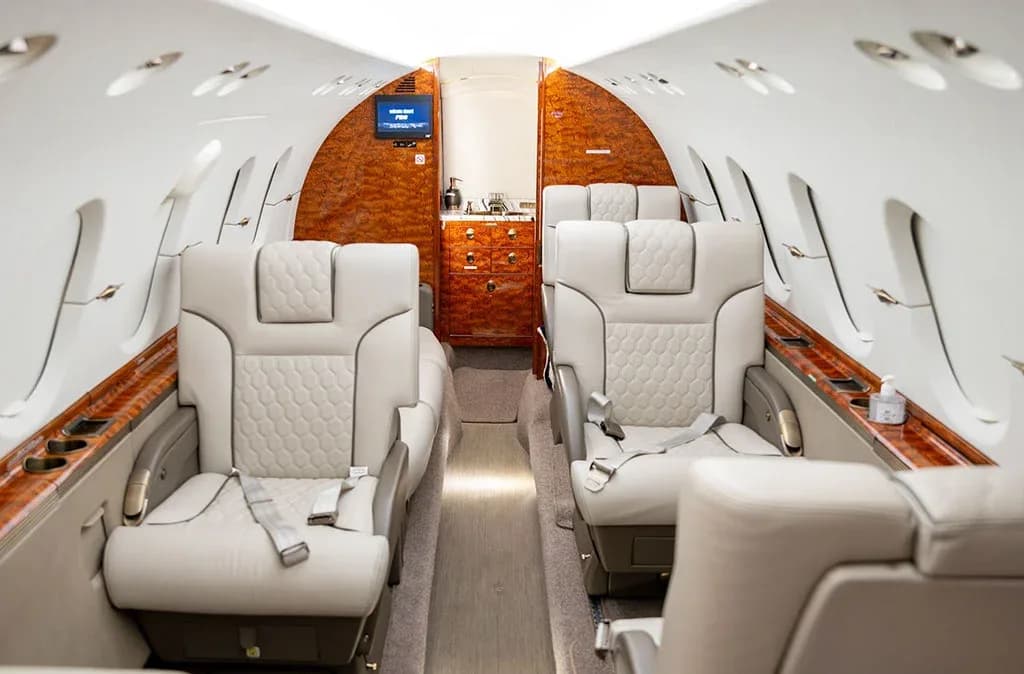 The luxurious interior of a private jet cabin with light grey quilted leather seats and polished wood accents.