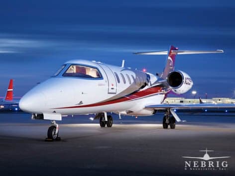 A white business jet with red and purple accents is parked on an airport tarmac at dusk.
