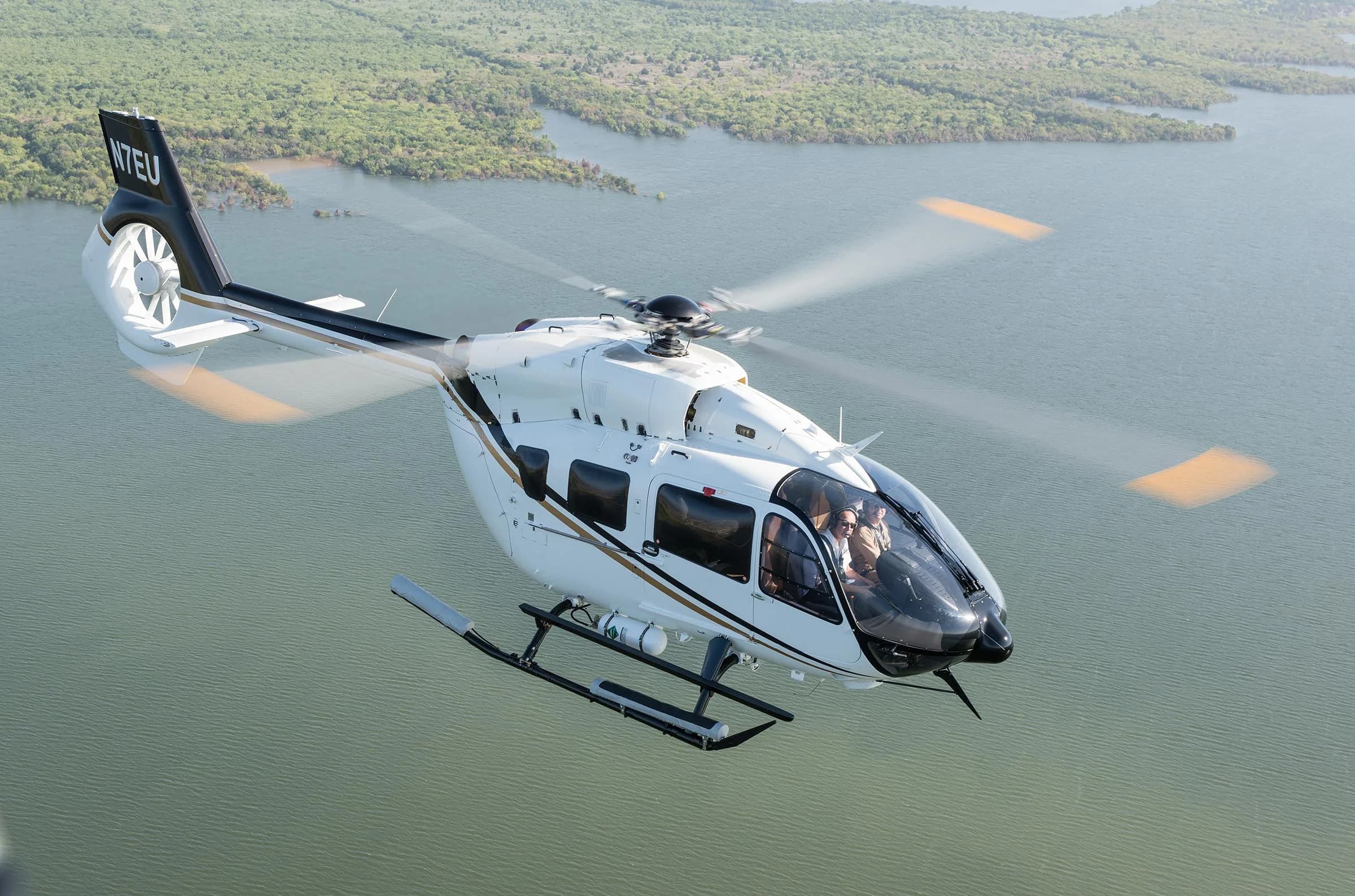 A white and gold Airbus EC145 helicopter in flight over a lake.