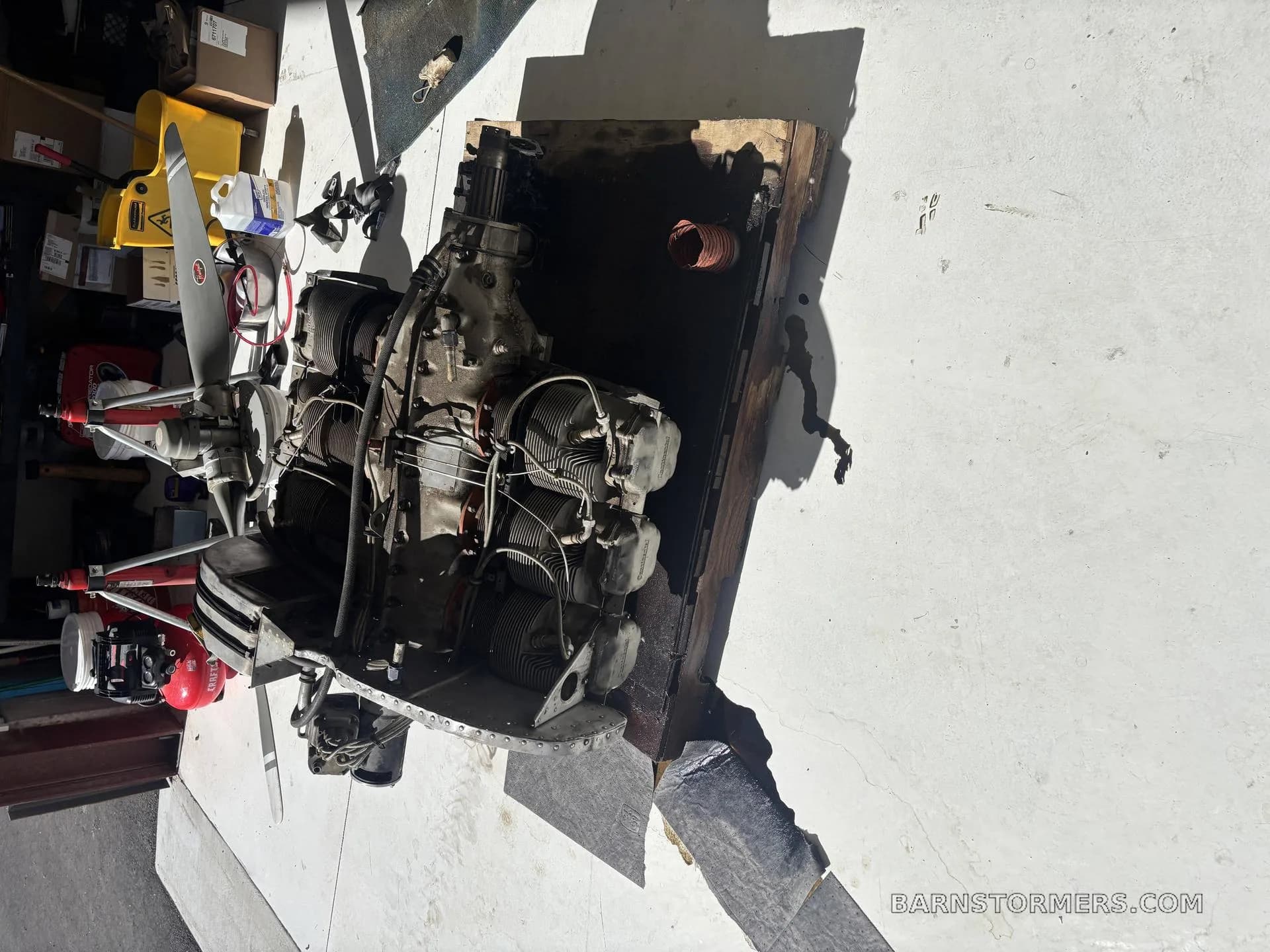 A top-down view of a disassembled aircraft engine with a propeller, mounted on a wooden stand in a sunlit garage.