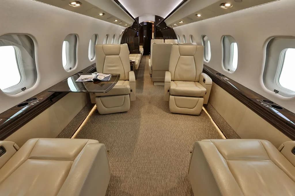 The luxurious cabin interior of a business jet, featuring beige leather seats, a table, and large windows.