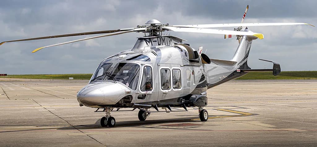 A silver and dark grey AgustaWestland AW169 helicopter (M-UMJR) parked on an airfield under a cloudy sky.