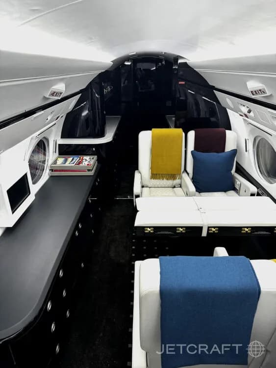 A view inside the private jet cabin showing white seats with blue and yellow throws, dark cabinetry, and a table.