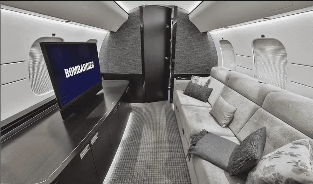 A private jet cabin interior with a long sofa, a TV displaying "BOMBARDIER", and dark textured walls.