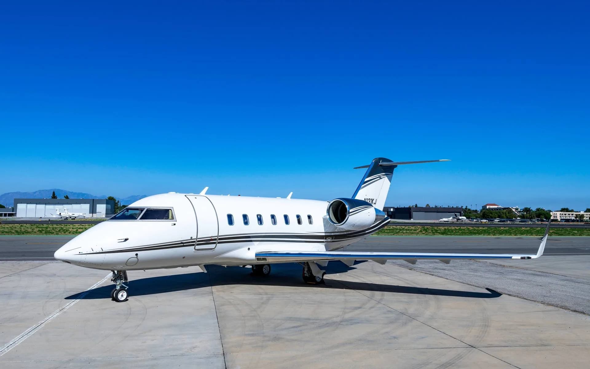 A white Bombardier Challenger business jet with dark and white stripes is parked on an airport tarmac under a clear blue sky.