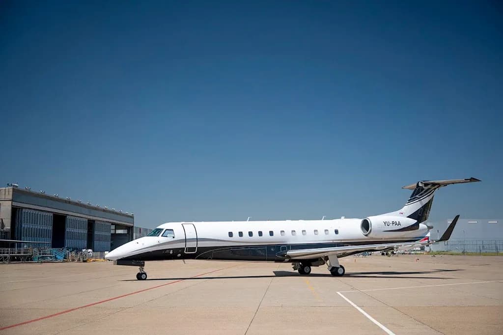 A white and black Embraer Legacy 600 business jet with registration YU-PAA parked on a tarmac next to a hangar.