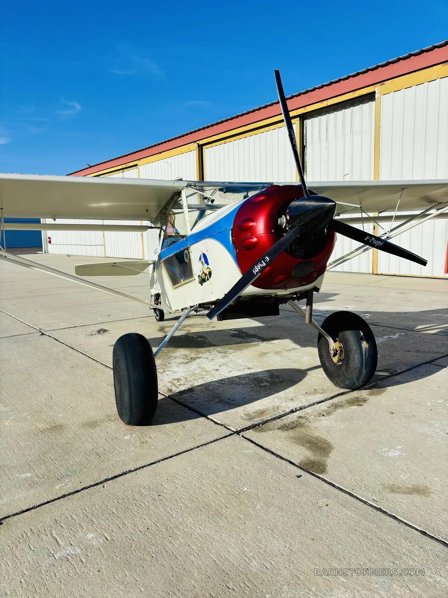 A white Kitfox aircraft with a red and blue cowl, large tires, parked on a tarmac in front of hangars.
