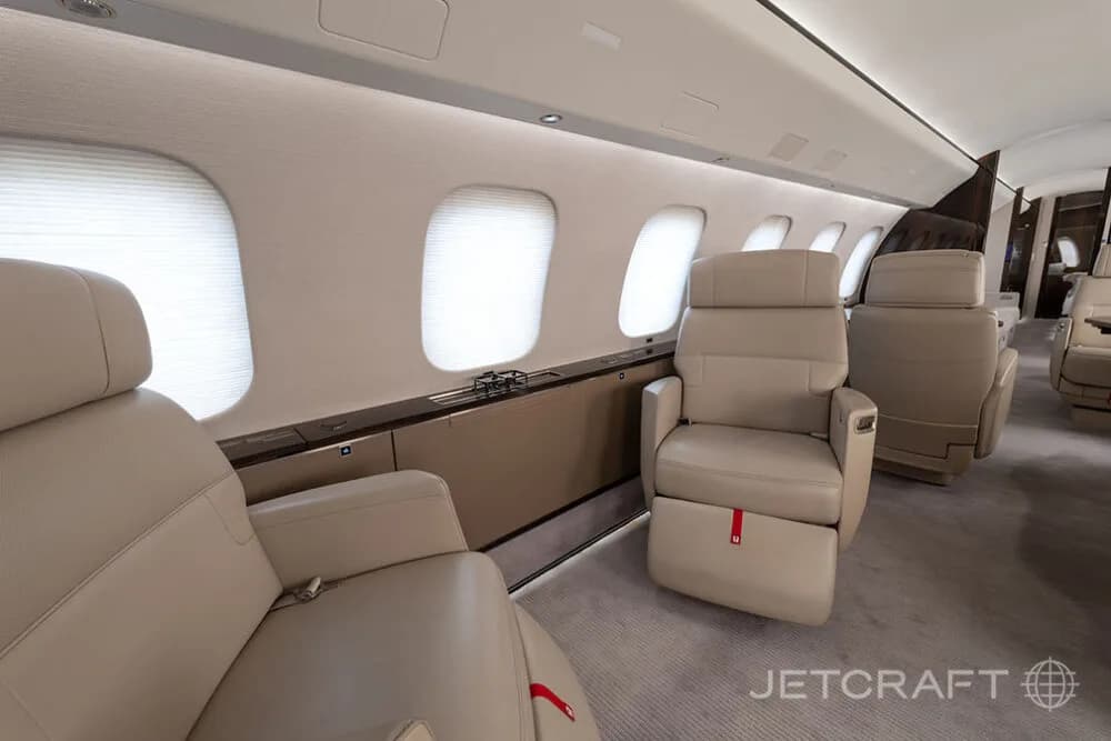 A view inside a private jet cabin, showing two light beige leather seats and multiple large windows with closed shades.