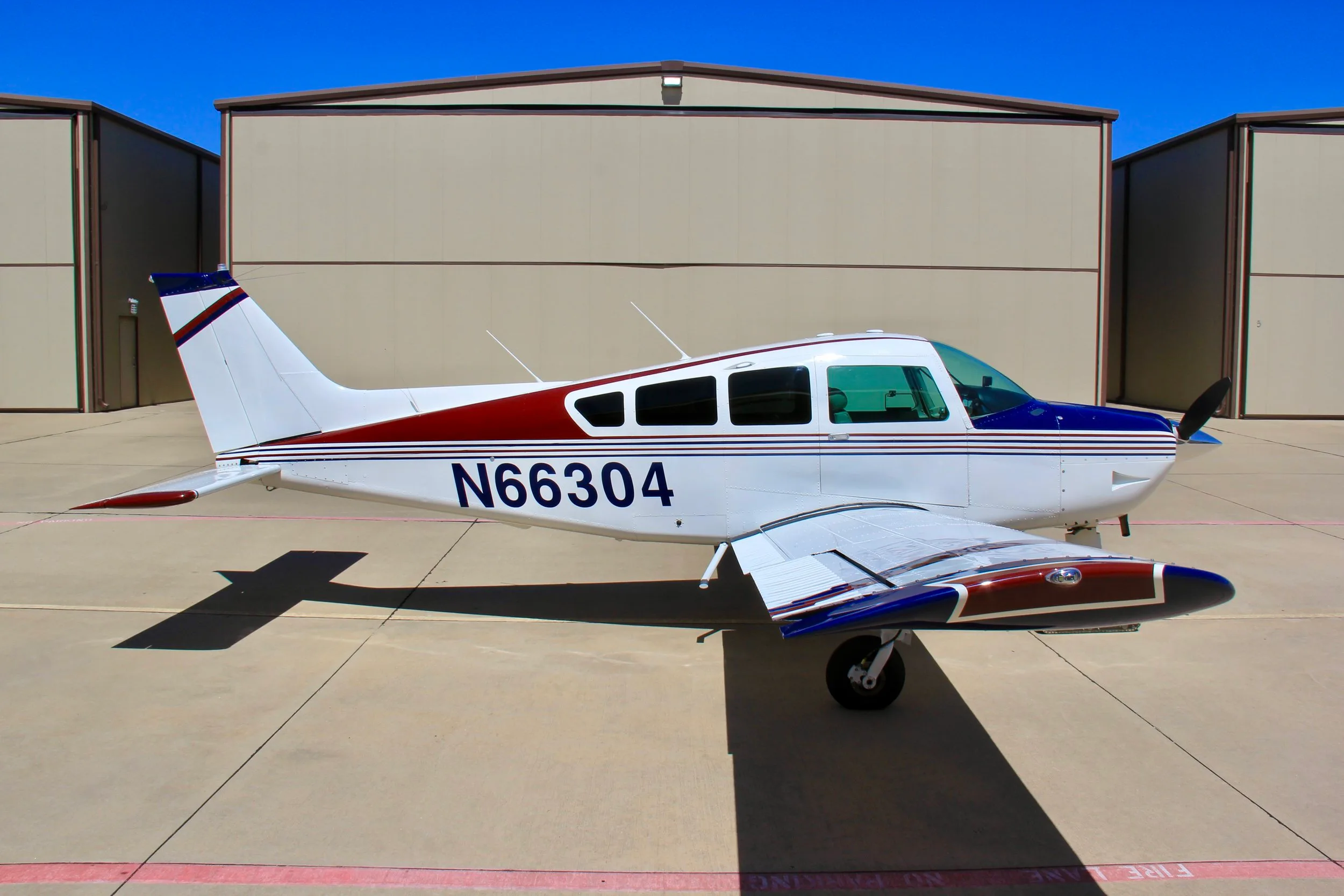 A side profile of the white, blue, and red single-engine aircraft (N66304) parked on an tarmac in front of hangars.