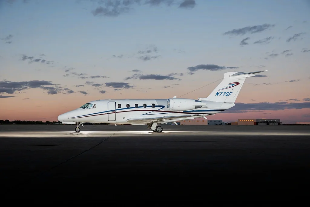 A white Cessna Citation business jet with blue and red stripes on the tail, parked on a tarmac at dusk.