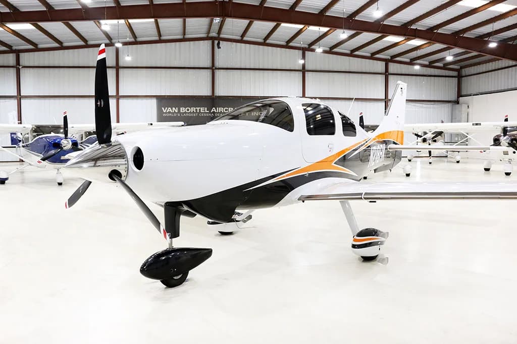 Front-left view of a white, black, and orange Cessna TTX aircraft with a propeller, inside a hangar.