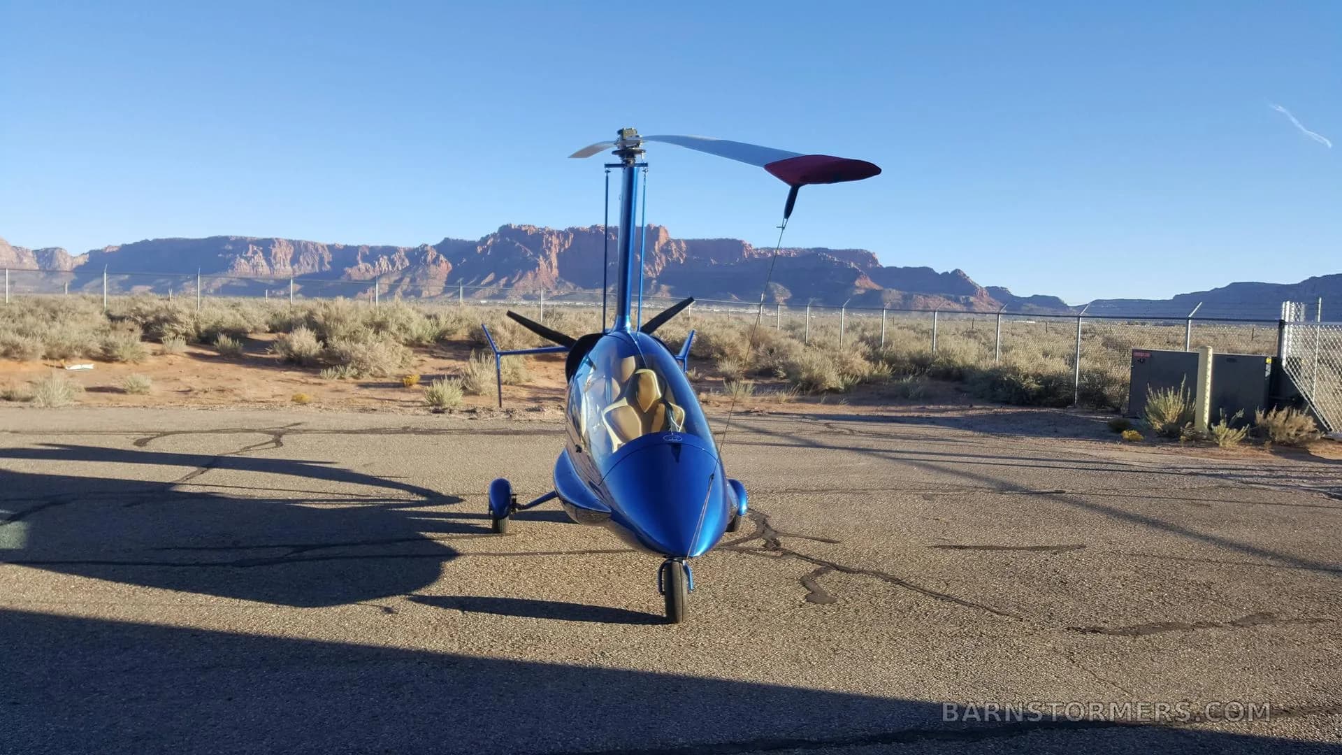 A blue enclosed gyrocopter is viewed from the front, parked on an asphalt lot with a desert and mountains in the background under a clear sky.