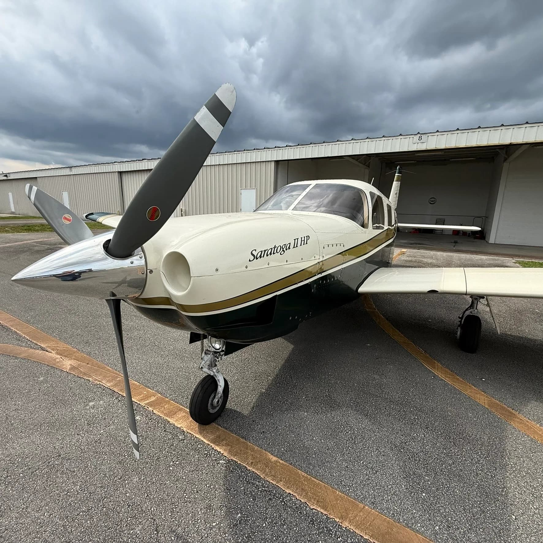 A Piper Saratoga II HP, N221AH, with a white, gold, and dark green paint scheme, parked on an airport tarmac under a cloudy sky.