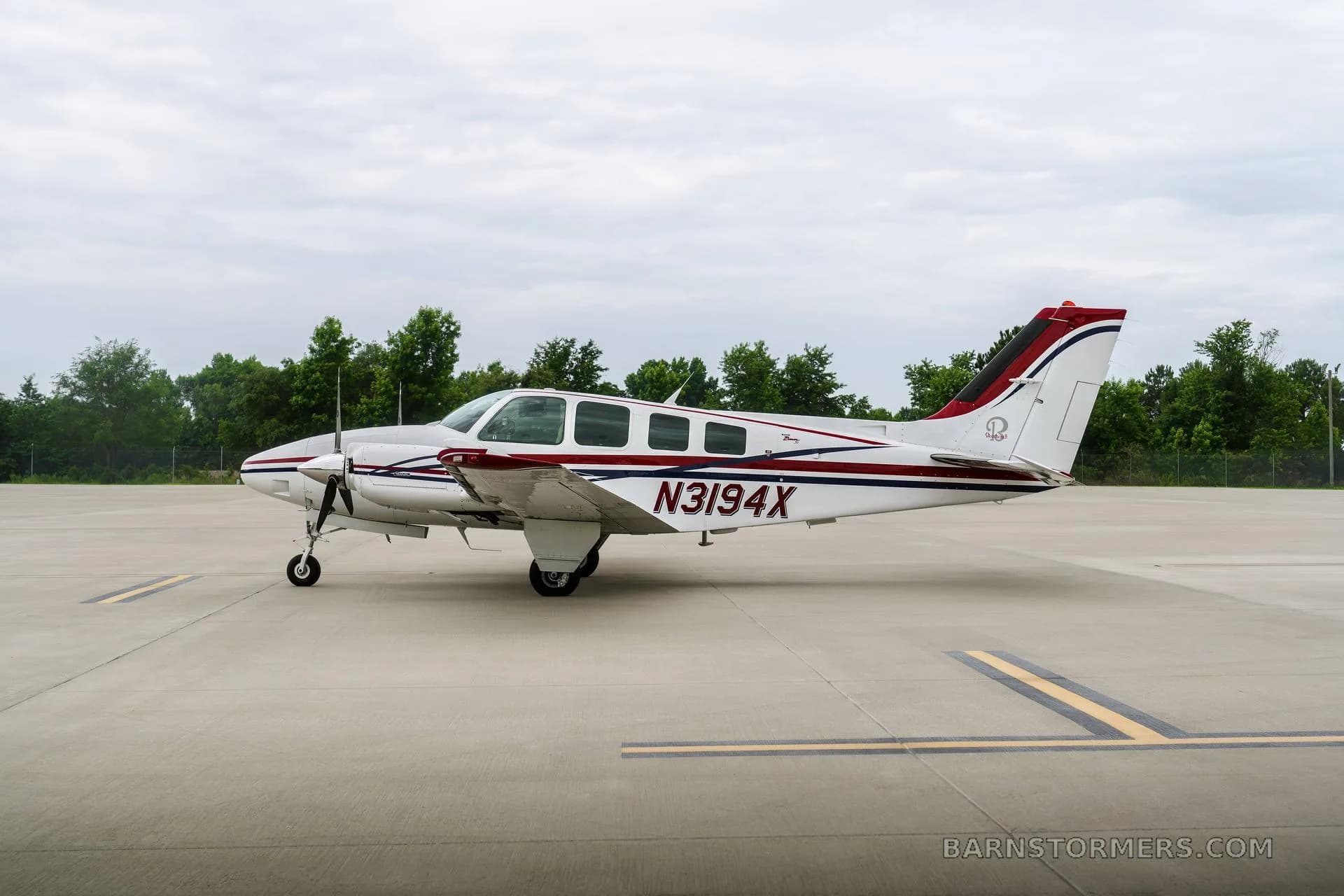 A side view of a white twin-engine propeller aircraft with red and blue stripes, registration N3194X, parked on an airfield with trees in the background.