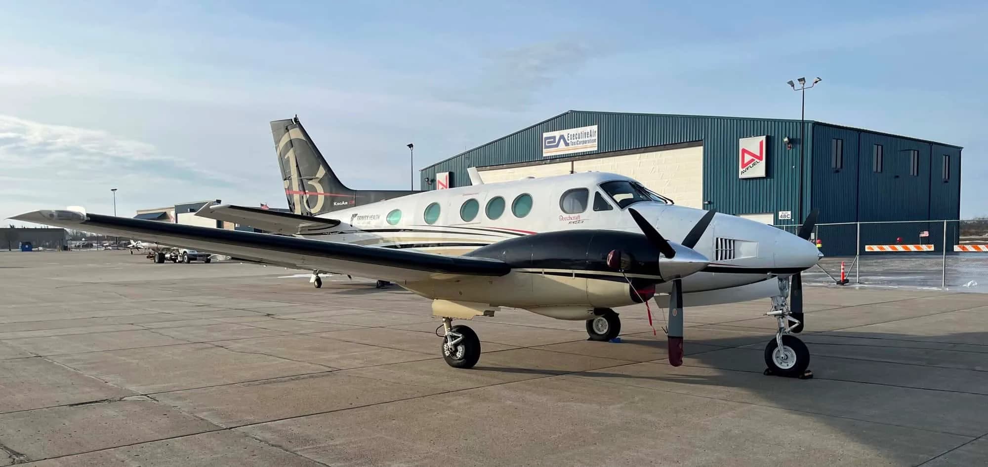 A white and black twin-engine turboprop aircraft with a distinctive tail logo, parked on an airfield in front of a hangar.