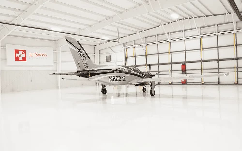 A Piper M600/SLS aircraft (N600AE) with a black and silver paint scheme is parked in a clean, white hangar, viewed from the front-left.