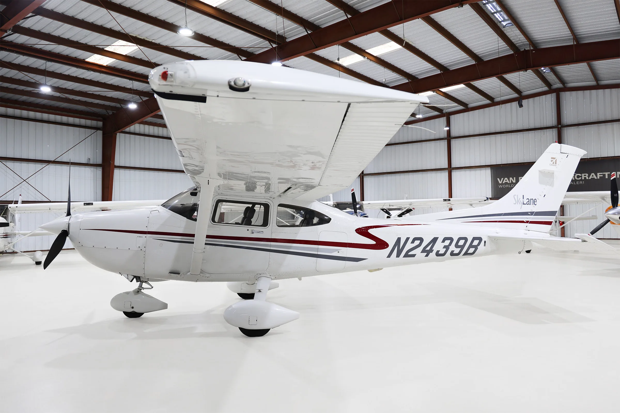 A white Cessna 182 Skylane with red and grey stripes, parked in a hangar, viewed from the side with the wing raised.