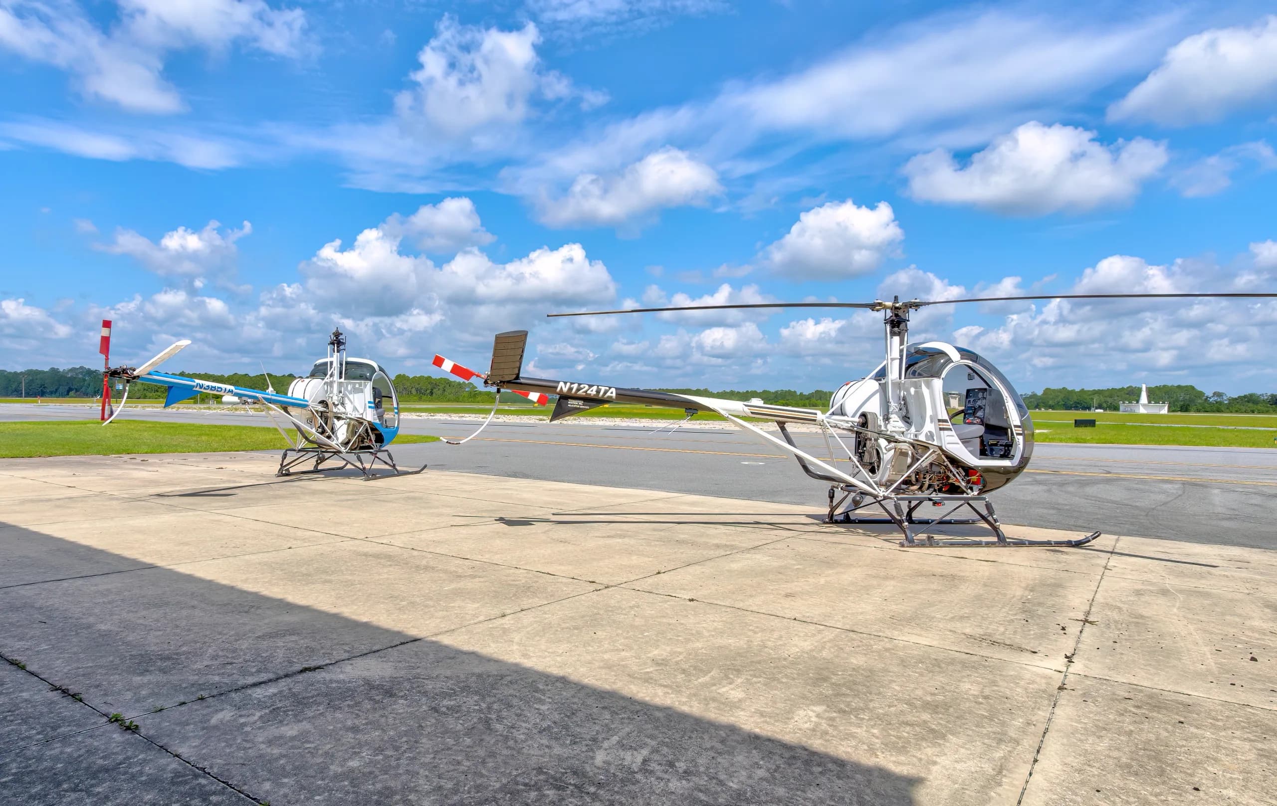 Two Schweizer 300CBi helicopters, N385TA (blue and white) and N124TA (black and white), parked side-by-side on an airfield tarmac.