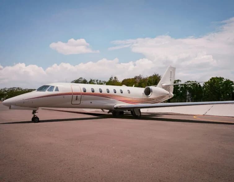A white private jet with red and gold stripes on its fuselage is parked on an asphalt apron under a partly cloudy sky.