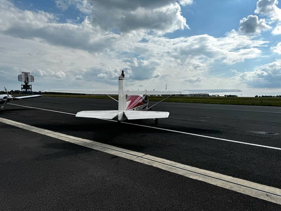 A white and red Cessna 150 G-ASMS parked on the tarmac, viewed from directly behind, with an airport beacon in the distance.