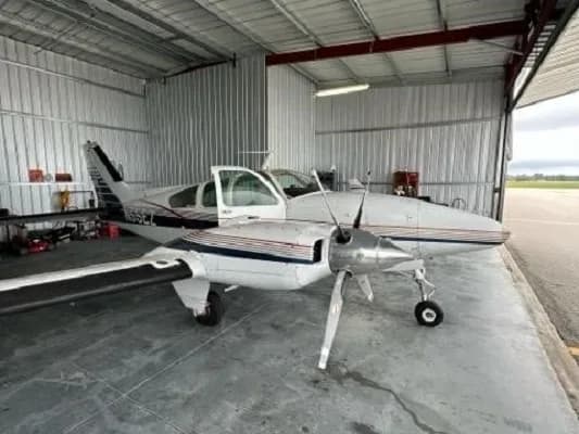 A white and blue twin-engine aircraft, N55EZ, is parked inside a hangar with the door open, viewed from the front-right.