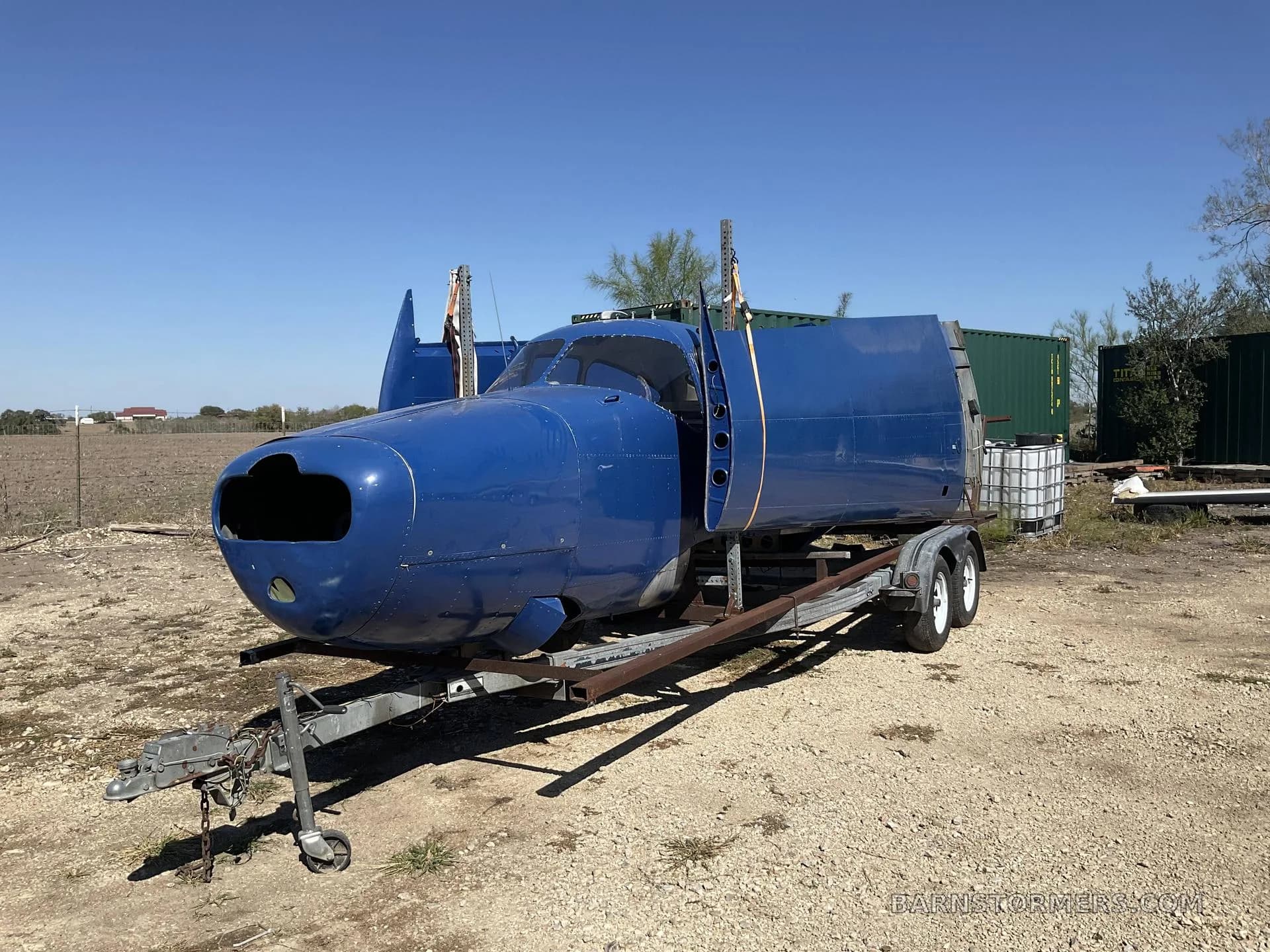 A blue aircraft fuselage, missing wings and engine, sits on a trailer in a gravel lot under a clear sky.