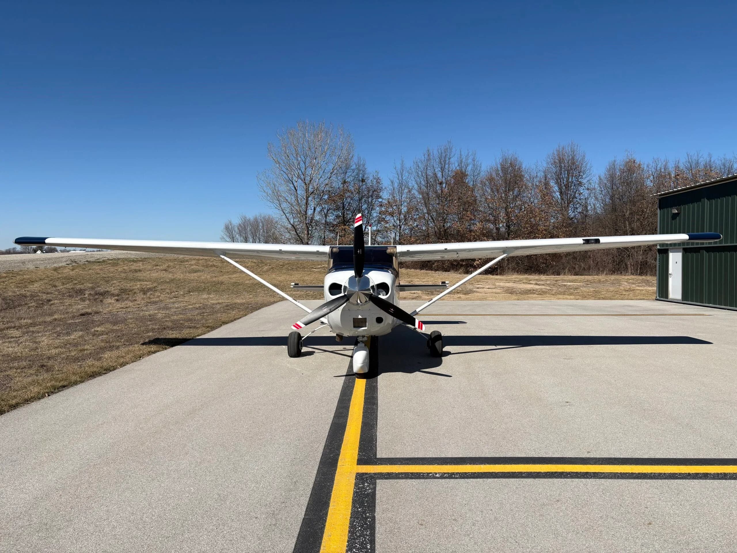 Front view of a white single-engine propeller aircraft with black and red propeller tips on an asphalt runway.