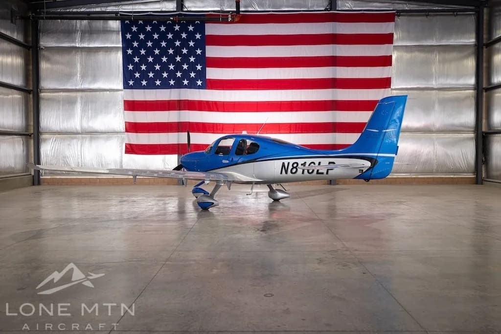 A blue and silver Cirrus SR22 aircraft, registration N816LP, parked in a hangar with an American flag in the background.