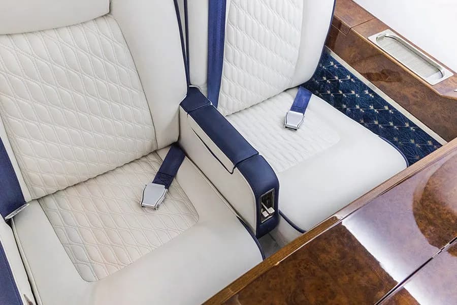 A close-up view of a white quilted leather seat with blue accents and a seatbelt in a private jet.