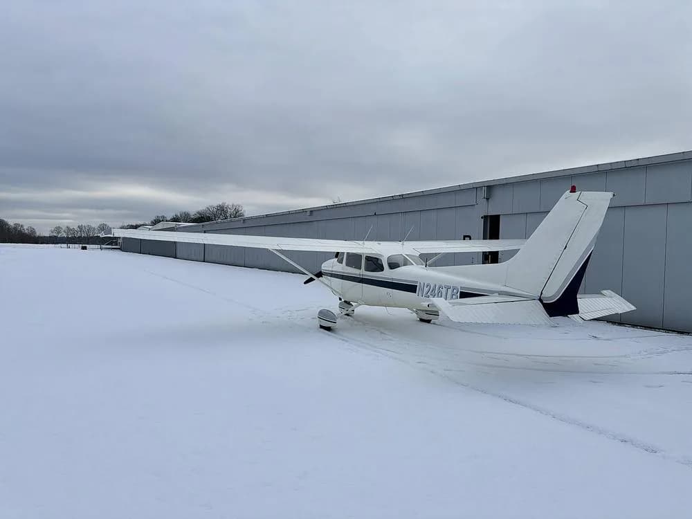 Rear-left view of a white Cessna 172 N246TB with dark blue tail stripes, parked in the snow next to a hangar.