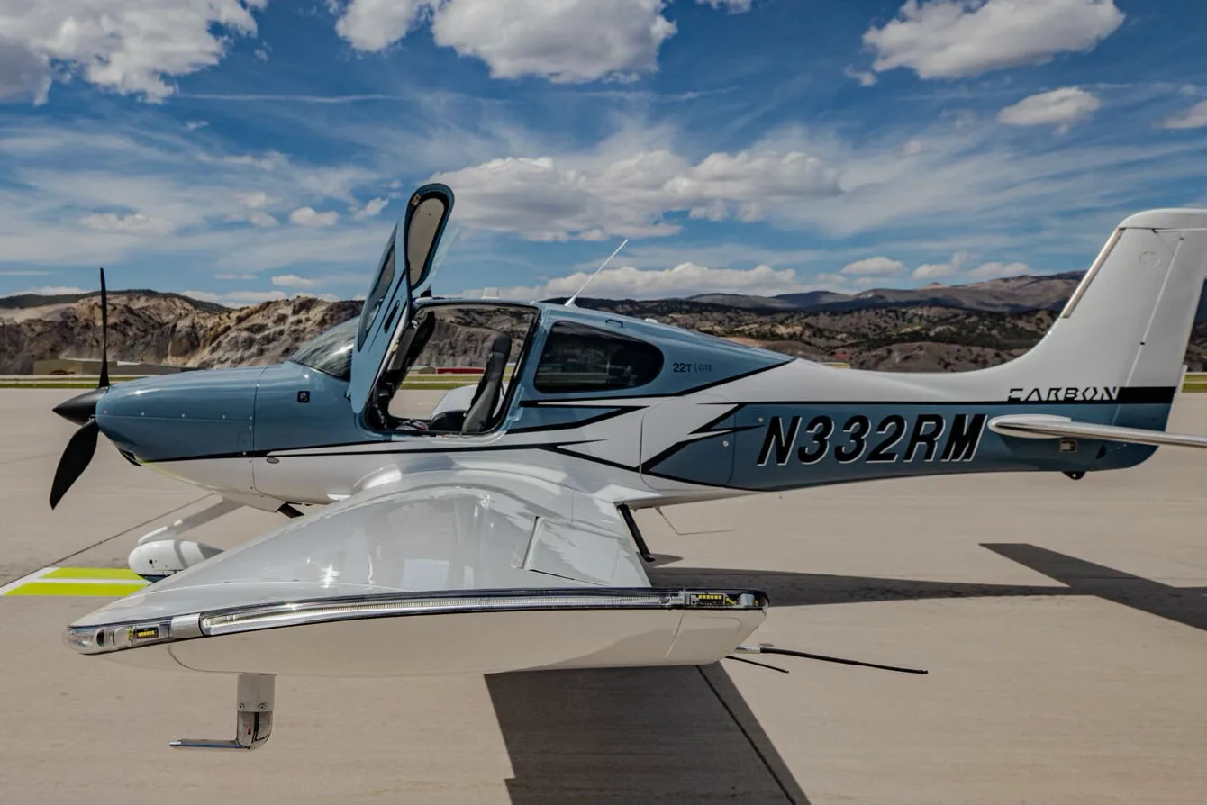Side view of a blue and white Cirrus SR22T GTS aircraft, registration N332RM, with its gull-wing doors open, parked on an airport tarmac.