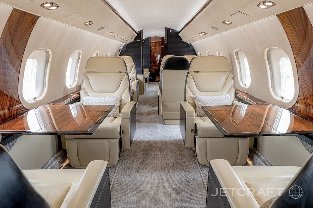 Forward view of the aircraft cabin with tan leather seats and wood trim, with tables extended.