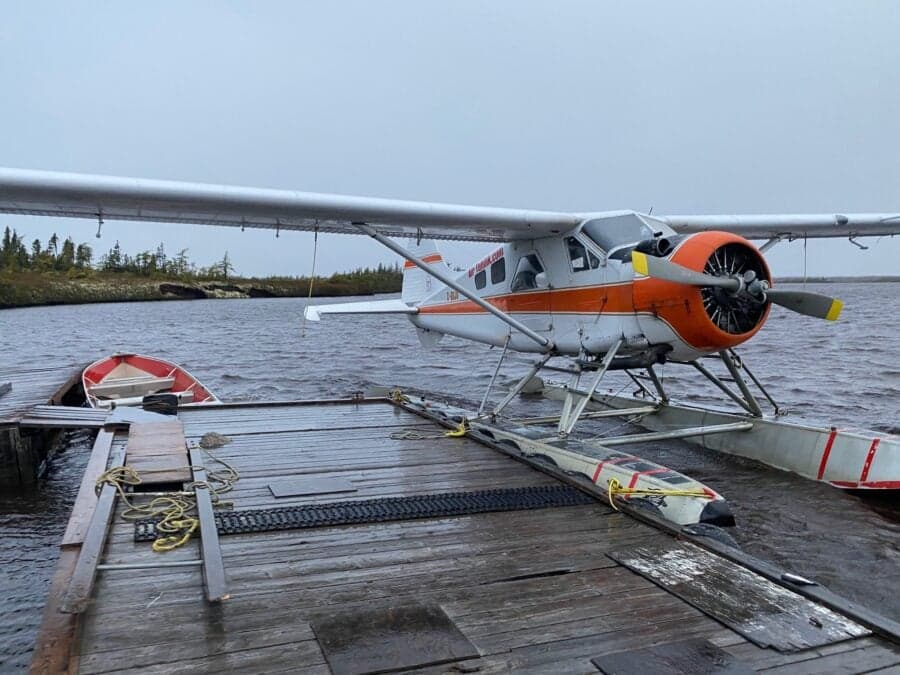 A de Havilland Beaver floatplane is docked on a wooden platform on a lake with a small boat nearby.