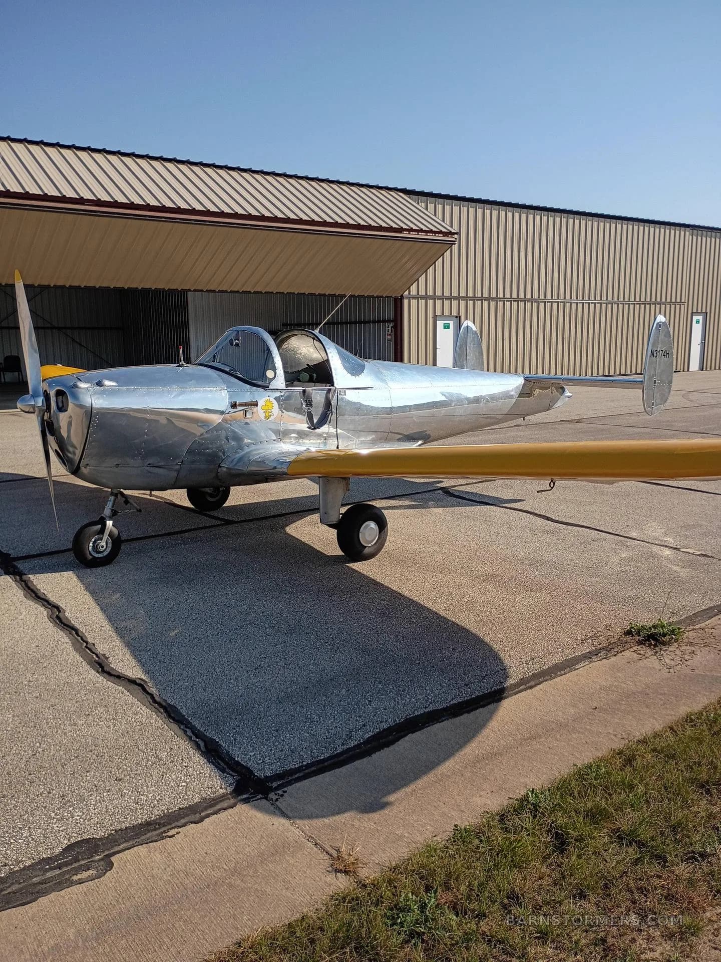 A shiny silver single-engine aircraft with yellow wings parked on an asphalt tarmac next to a hangar.