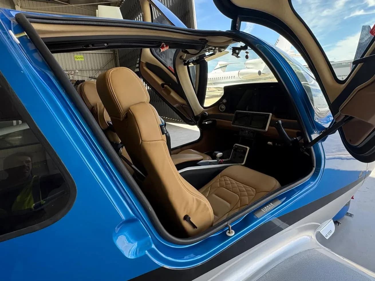 The tan leather interior and cockpit of a blue Cirrus SR22 aircraft with its door open, viewed from the right side.