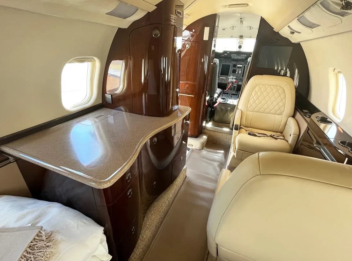 The business jet cabin features cream leather seats, dark wood cabinetry with a light countertop, and a view towards the cockpit.