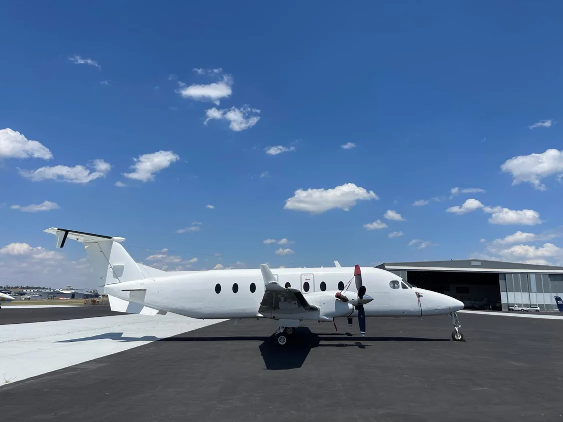 A white twin-turboprop aircraft with red propeller tips parked on an asphalt tarmac under a blue sky with scattered clouds.