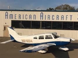 A white and blue Piper PA-28-180 Cherokee, registration N61884, parked on an apron in front of an 'AMERICAN AIRCRAFT' building.