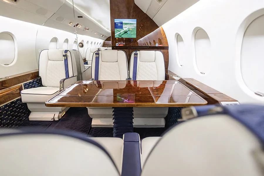 A private jet cabin with white and blue seats facing a large polished wooden table.