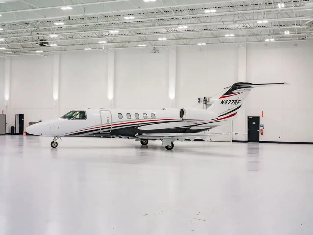 A white Cessna Citation Latitude business jet with black, red, and grey stripes is parked in a brightly lit hangar.