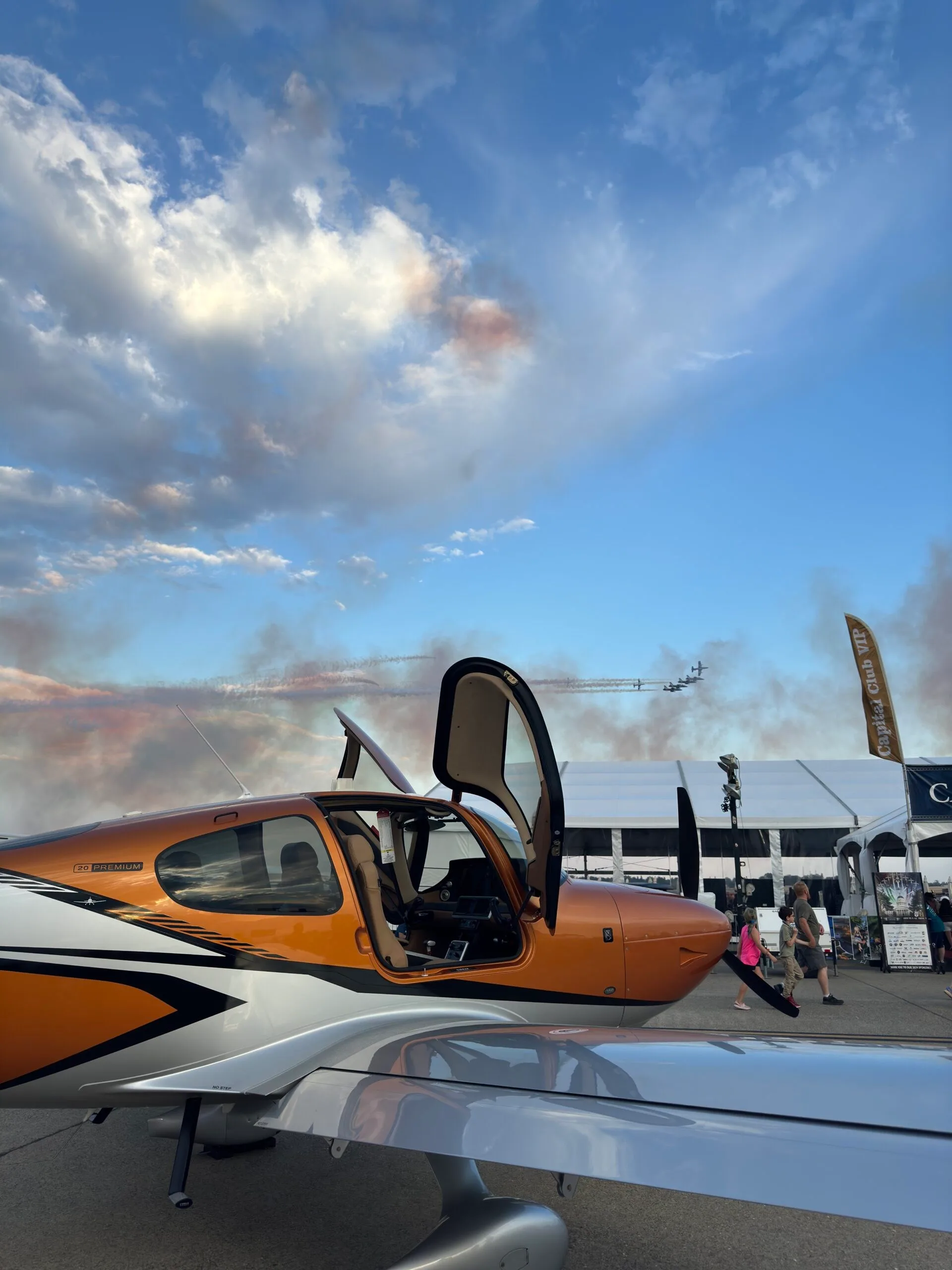 A Cirrus SR22 aircraft with orange and silver paint, door open, on an airfield with an airshow in the background.