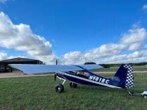 A blue and white ACA aircraft, registration N101BC, parked on a grassy field with a hangar in the background.