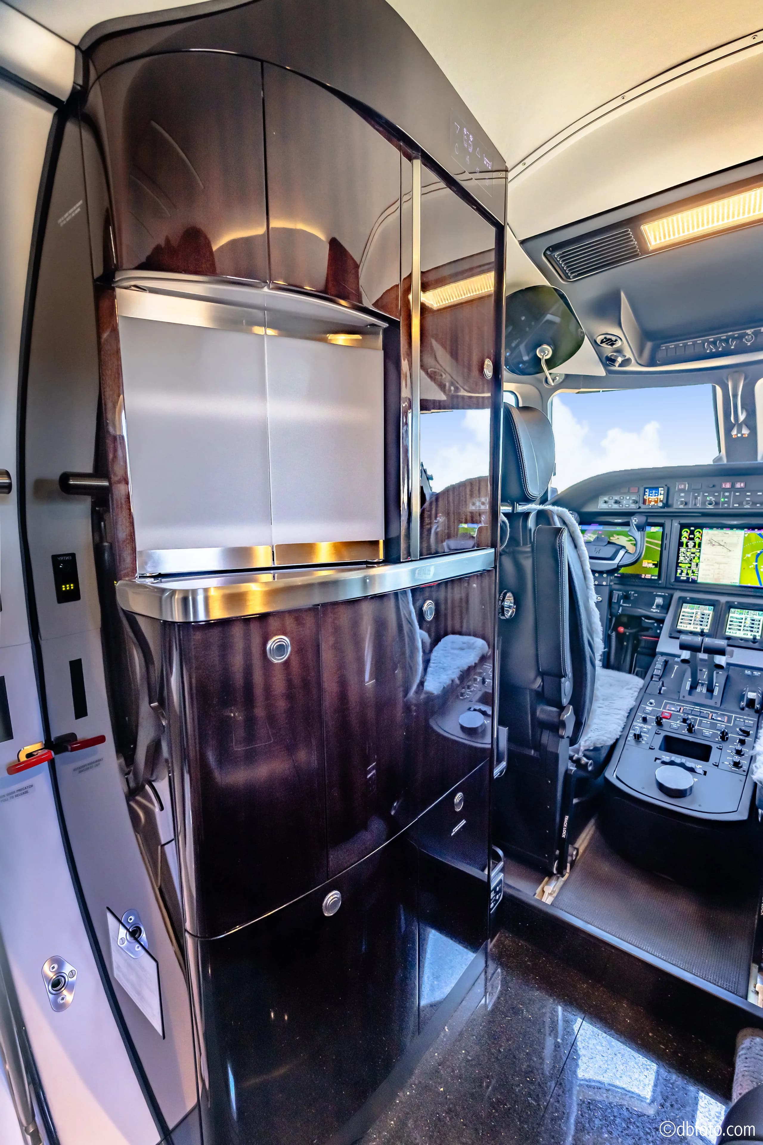 The business jet's galley area with dark wood cabinetry and a view into the cockpit.