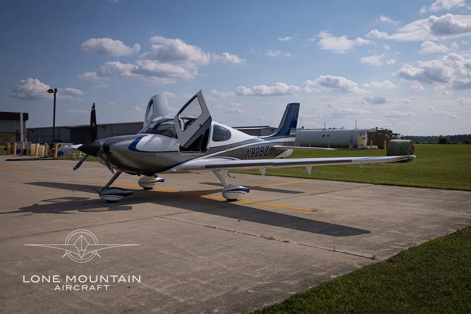 A three-quarter front view of the white and silver Cirrus SR22 aircraft N92BZ with blue accents, parked on an airfield with its doors open, with buildings in the background.