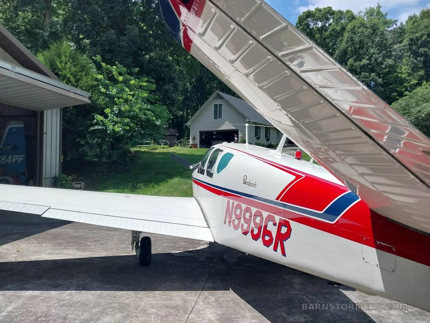 A white Beechcraft Bonanza with red and blue stripes, registration N999CR, viewed from the rear-right, parked next to a hangar.