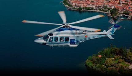 A silver and blue helicopter flies over a body of water near a town.
