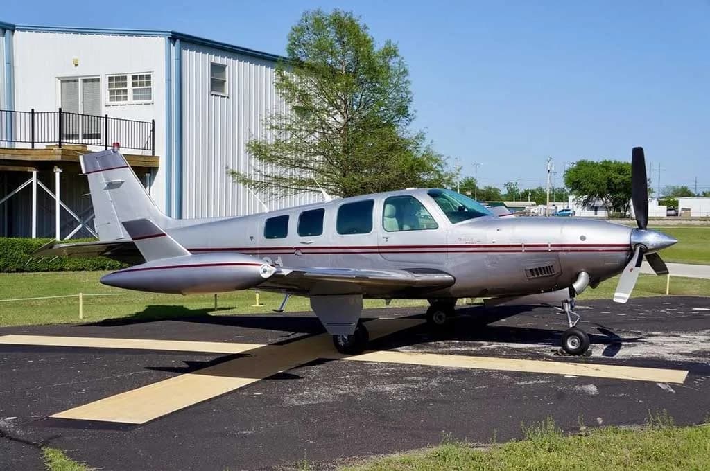 A silver and red Beechcraft Baron 58 is parked on an asphalt ramp next to a white hangar.